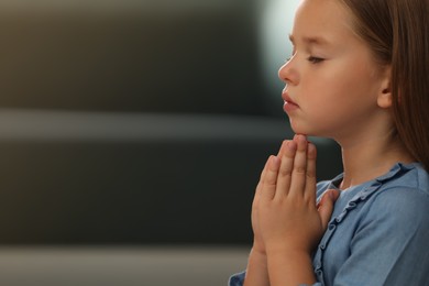 Cute little girl with hands clasped together praying on blurred background. Space for text