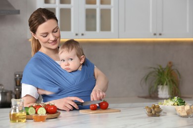 Photo of Mother cutting tomatoes while holding her child in sling (baby carrier) in kitchen