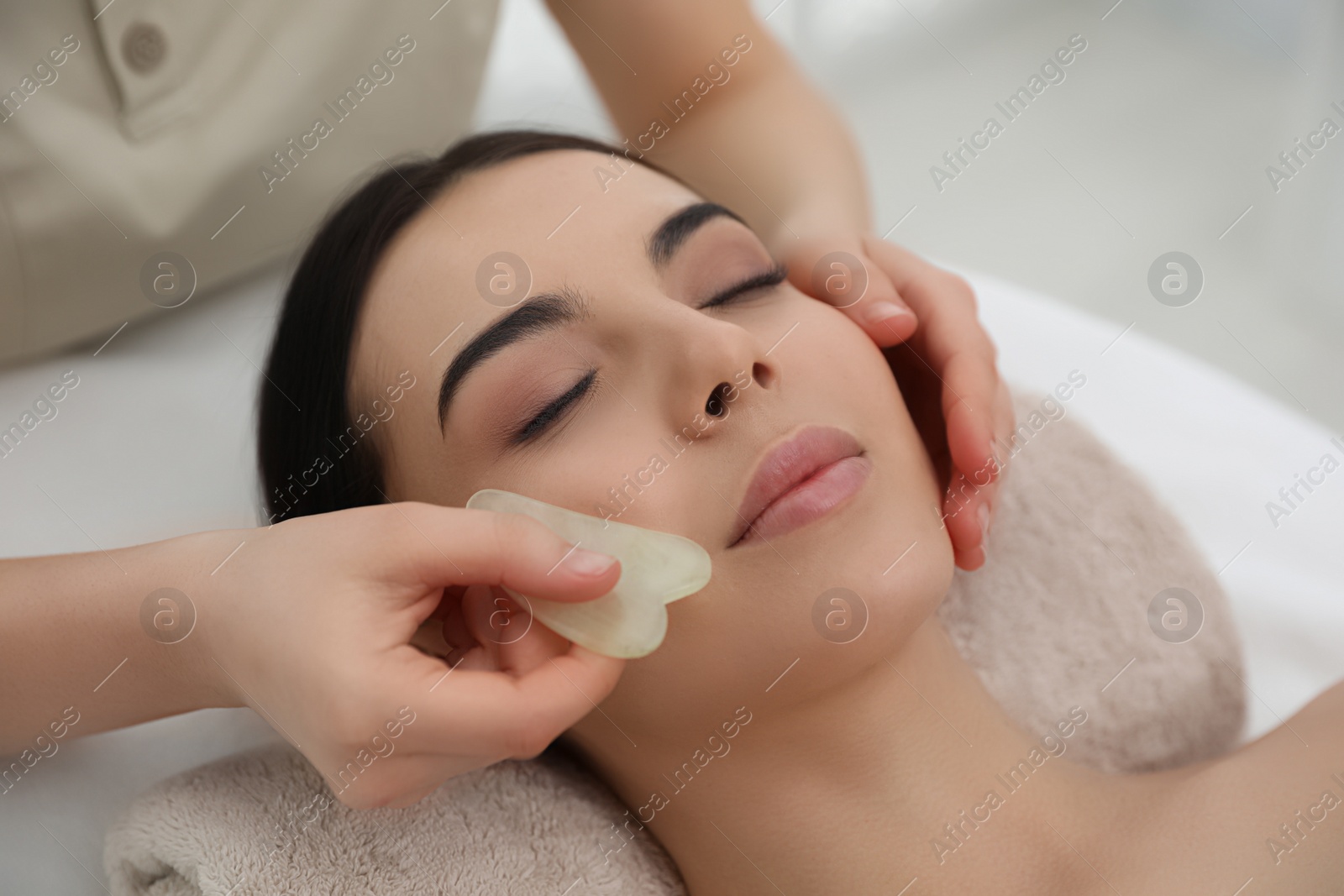 Photo of Young woman receiving facial massage with gua sha tool in beauty salon