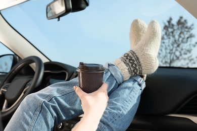 Photo of Young woman in warm socks holding her legs on car dashboard and drinking coffee. Cozy atmosphere