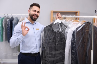 Photo of Dry-cleaning service. Happy worker holding hanger with denim jacket in plastic bag and showing ok gesture near rack with clothes indoors