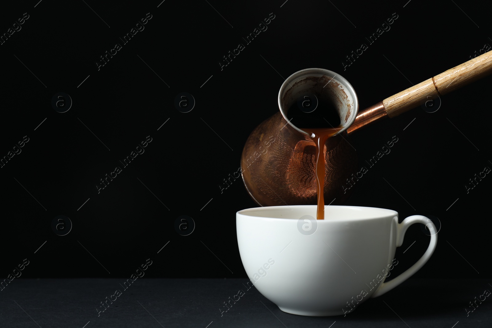 Photo of Turkish coffee. Pouring brewed beverage from cezve into cup on table against black background, space for text