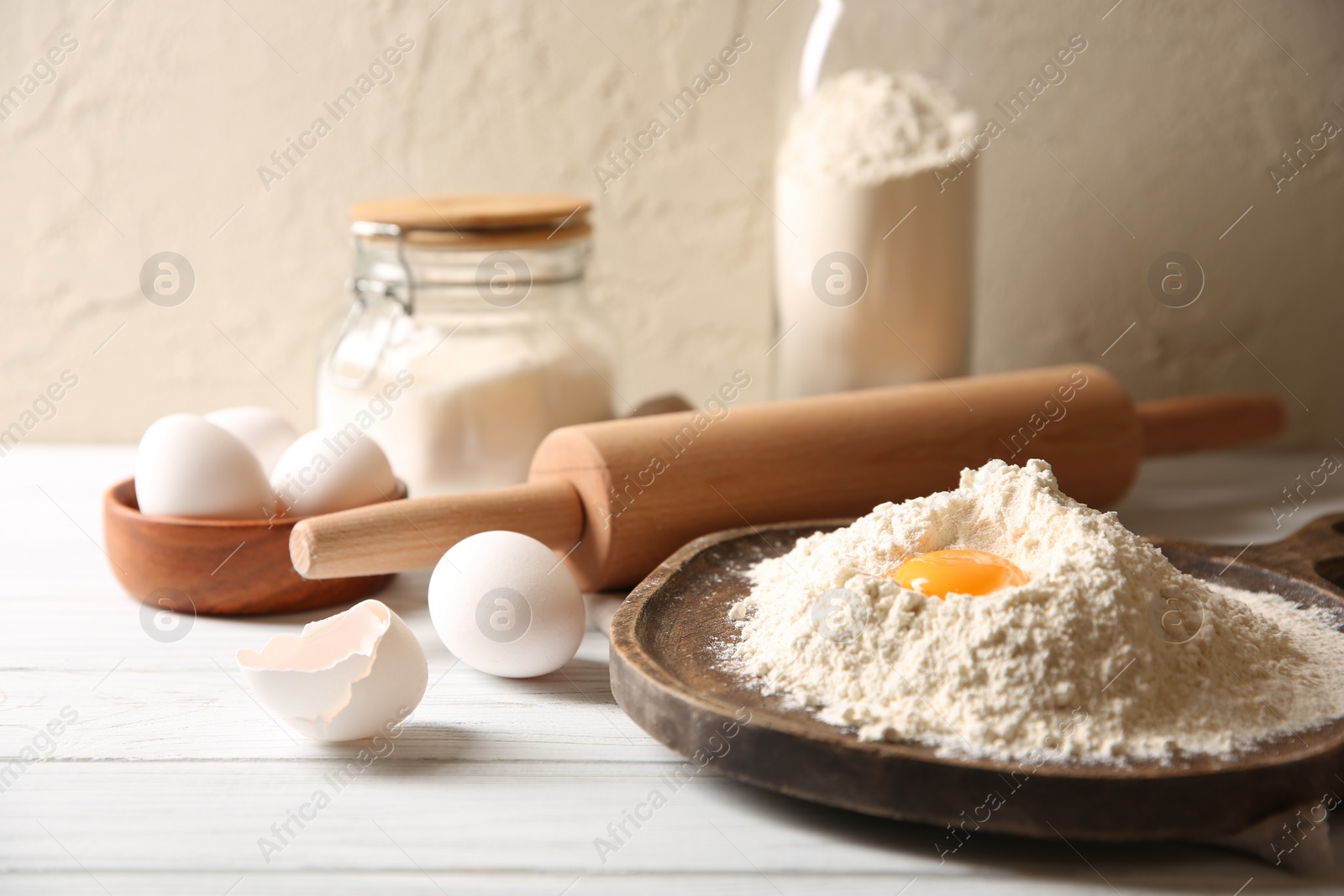 Photo of Making dough. Pile of flour with yolk, rolling pin and eggs on white wooden table, closeup