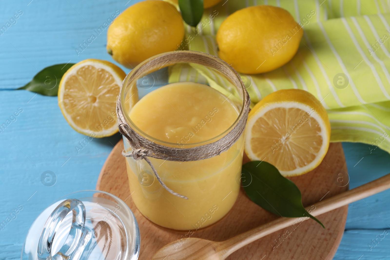 Photo of Delicious lemon curd in glass jar, fresh citrus fruits, spoon and green leaves on light blue wooden table