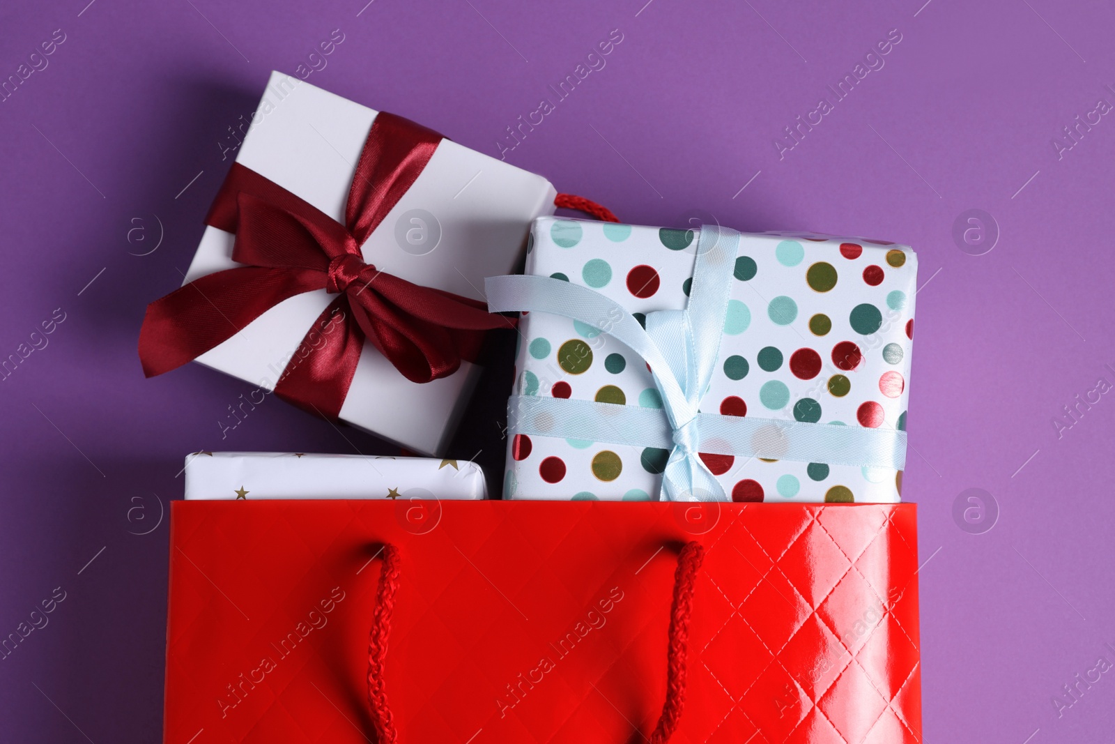 Photo of Red paper shopping bag full of gift boxes on purple background, flat lay