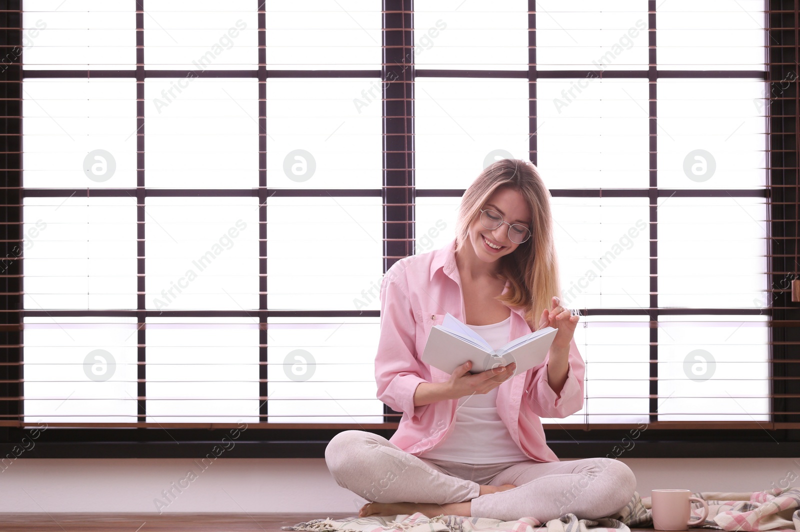 Photo of Young woman reading book near window with blinds at home. Space for text