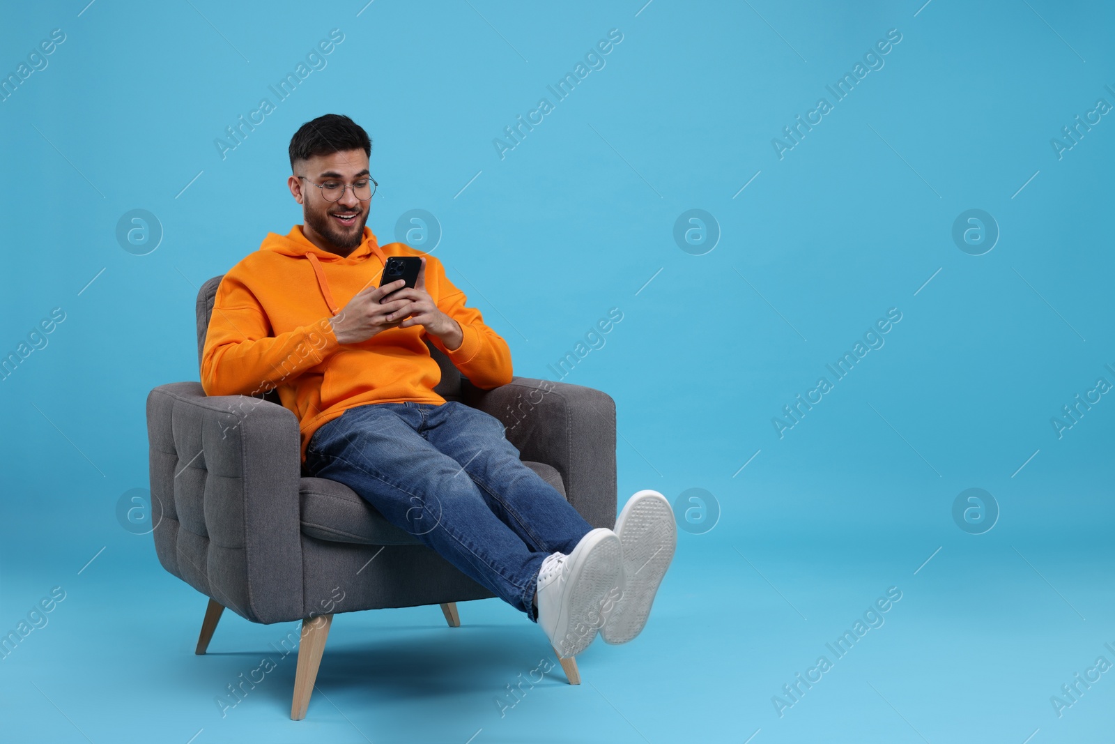 Photo of Happy young man using smartphone on armchair against light blue background, space for text