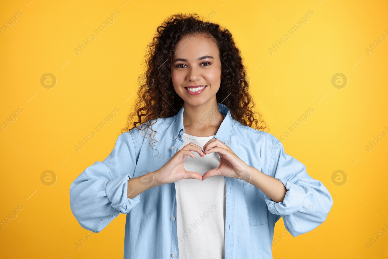 Photo of Happy young African-American woman making heart with hands on yellow background