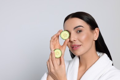 Woman in bathrobe holding pieces of cucumber on light grey background, space for text. Spa treatment