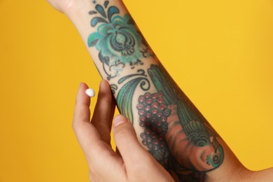 Photo of Woman applying cream on her arm with tattoos against yellow background, closeup