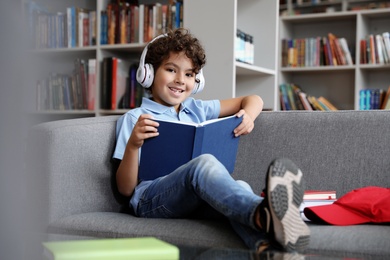Photo of Cute little boy in headphones reading book on sofa in library