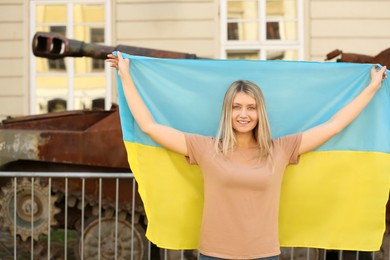 Photo of Happy woman holding Ukrainian flag near broken tank in city