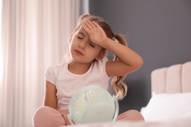 Photo of Little girl enjoying air flow from portable fan in room. Summer heat