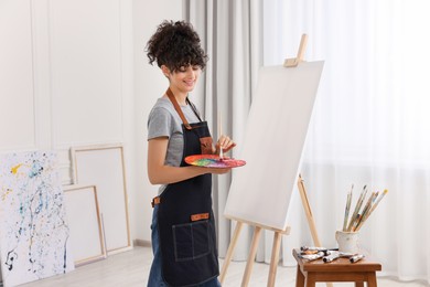Photo of Young woman mixing paints on palette with brush near easel in studio