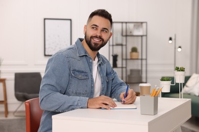 Photo of Young man writing in notebook at white table indoors