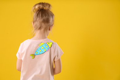 Photo of Little girl with paper fish on back against yellow background, space for text. April fool's day