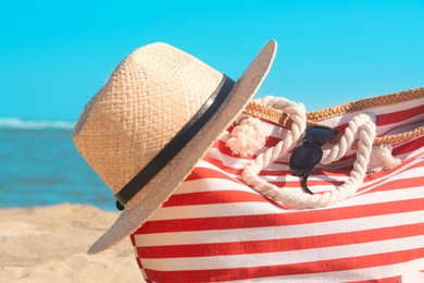Photo of Stylish striped bag with straw hat and sunglasses on sandy beach near sea, closeup