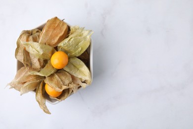 Photo of Ripe physalis fruits with calyxes in bowl on white marble table, top view. Space for text