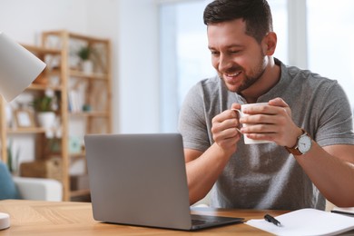 Photo of Happy man with cup of drink working on laptop at wooden desk in room