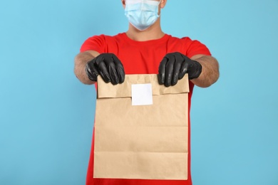 Photo of Courier in medical mask holding paper bag with takeaway food on light blue background, closeup. Delivery service during quarantine due to Covid-19 outbreak