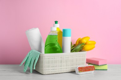 Plastic basket with different cleaning supplies and beautiful spring flowers on white wooden table against light pink background