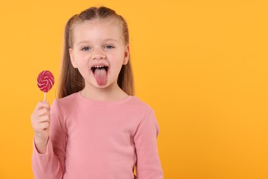 Photo of Funny little girl with bright lollipop swirl showing tongue on orange background, space for text