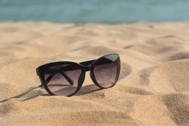 Photo of Stylish sunglasses on sandy beach near sea, closeup