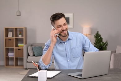 Photo of Young man talking on phone while working with laptop at desk in home office