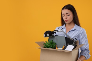 Photo of Unemployed problem. Sad woman with box of personal office belongings on orange background, space for text