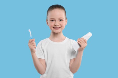 Photo of Happy girl holding toothbrush and tube of toothpaste on light blue background