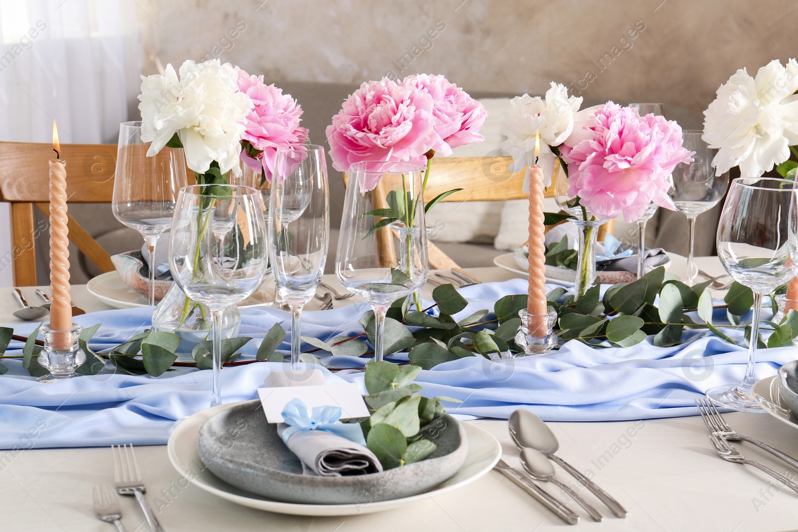 Photo of Beautiful table setting. Plate with greeting card, napkin and branch near glasses, peonies, burning candles and cutlery on table indoors