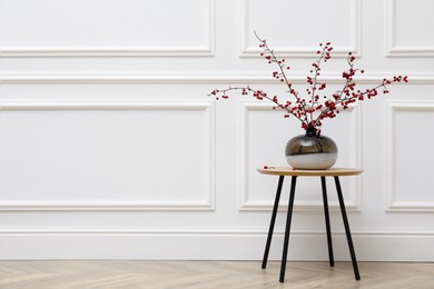 Photo of Hawthorn branches with red berries in vase on wooden table near white wall indoors, space for text