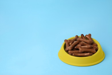 Photo of Yellow bowl with bone shaped dog cookies on light blue background, space for text
