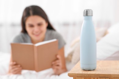 Photo of Young woman reading book in bedroom, focus on thermo bottle