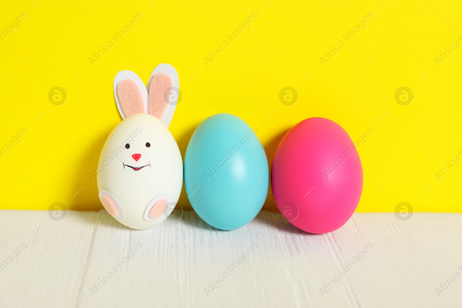 Photo of Bright Easter eggs and white one as cute bunny on wooden table against yellow background