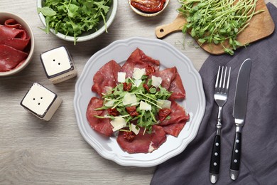 Plate of tasty bresaola salad with sun-dried tomatoes, parmesan cheese and cutlery on wooden table, flat lay