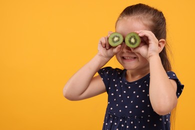 Photo of Smiling girl covering eyes with halves of fresh kiwi on orange background, space for text