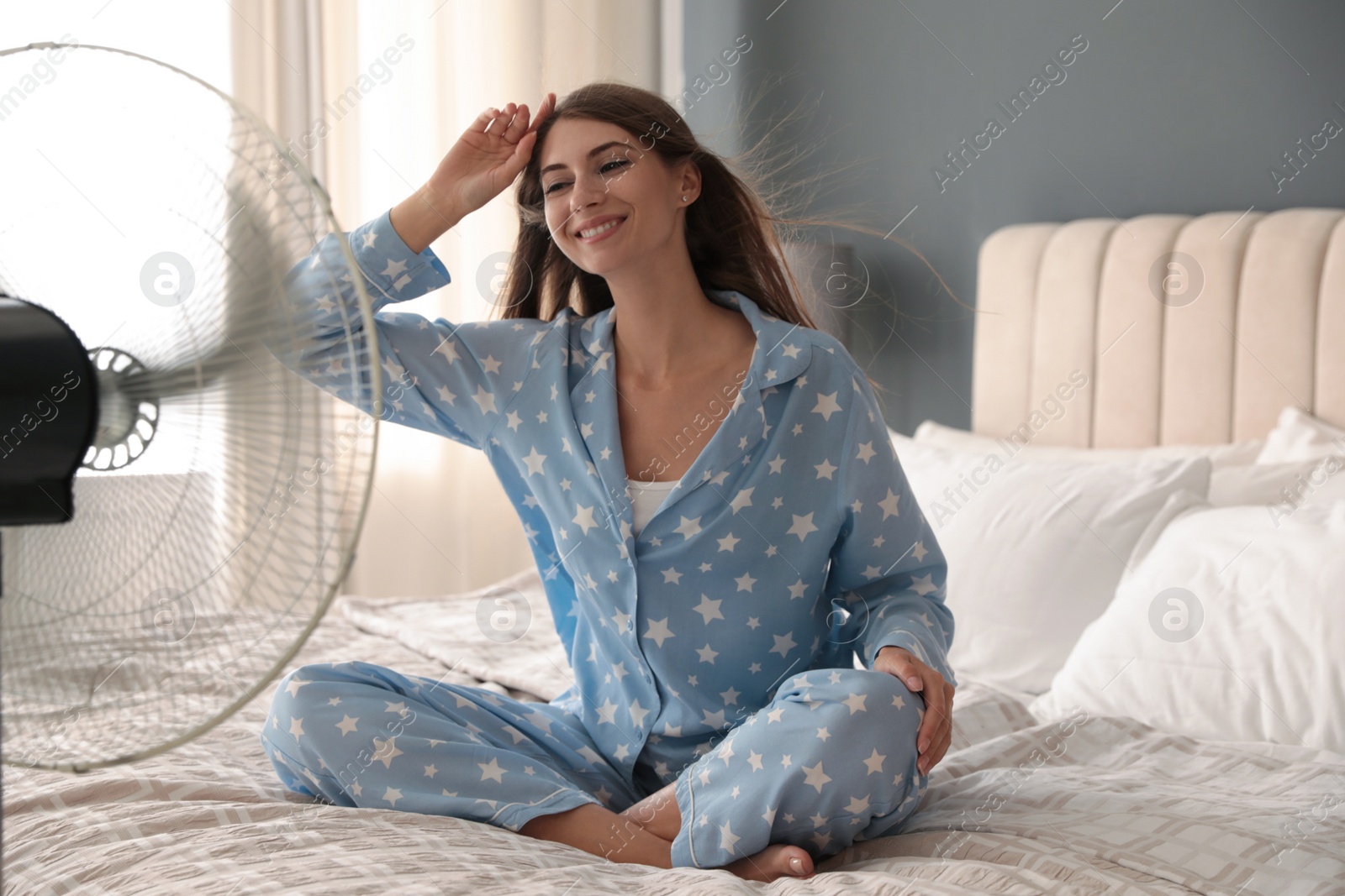 Photo of Woman enjoying air flow from fan on bed in room. Summer heat