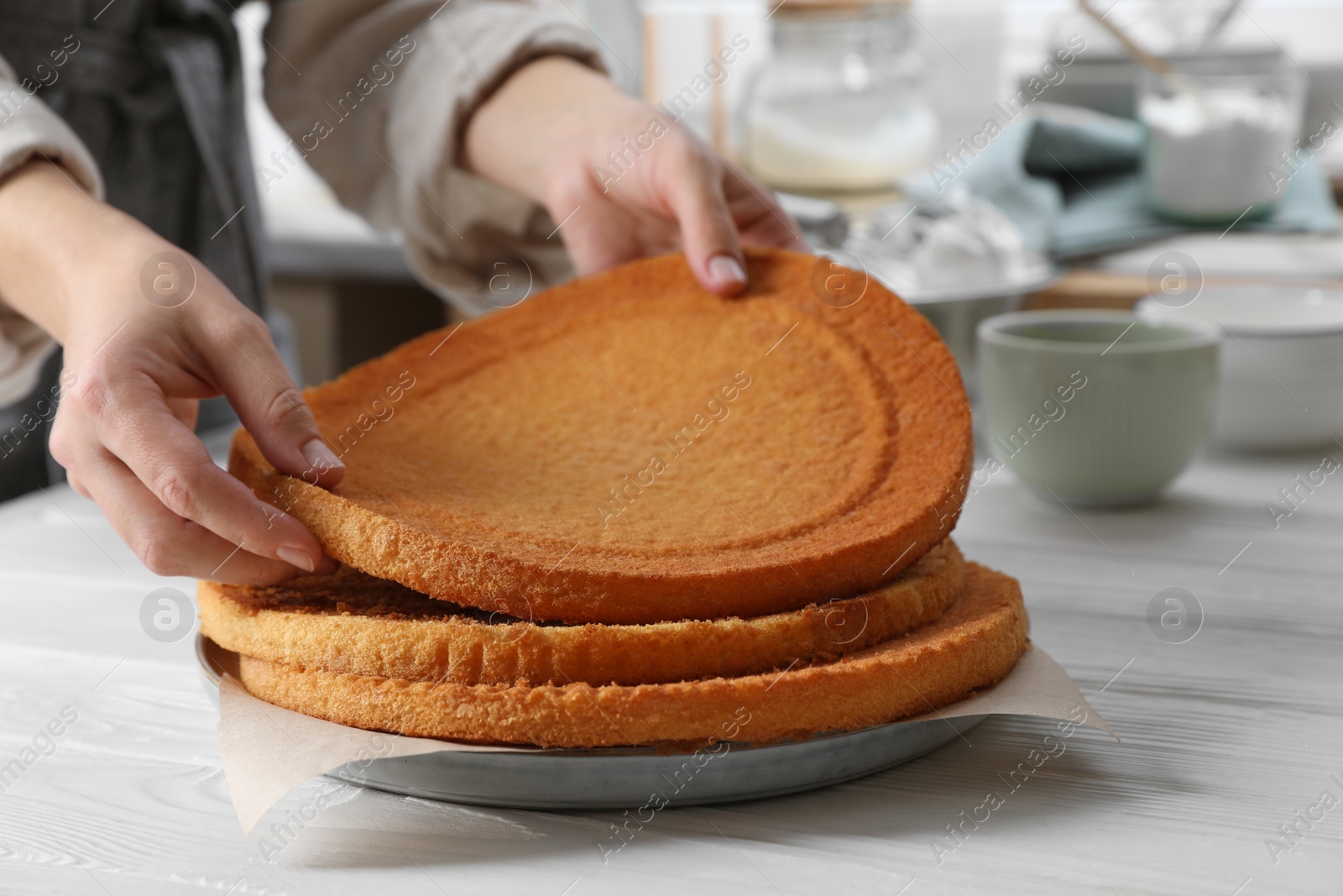 Photo of Woman stacking homemade sponge cakes at white wooden table in kitchen, closeup