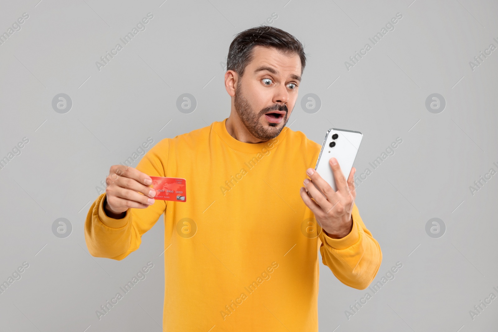 Photo of Shocked man with credit card and smartphone on grey background. Debt problem