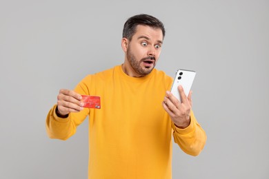 Shocked man with credit card and smartphone on grey background. Debt problem