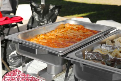Photo of Delicious stuffed cabbage, dumplings and patties in warmers on table outdoors. Volunteer food distribution