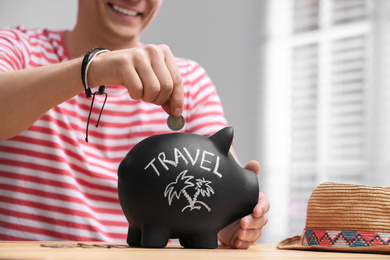 Photo of Man putting coin into piggy bank with word TRAVEL at table, closeup
