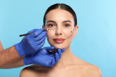 Image of Woman preparing for cosmetic surgery, light blue background. Doctor drawing markings on her face, closeup