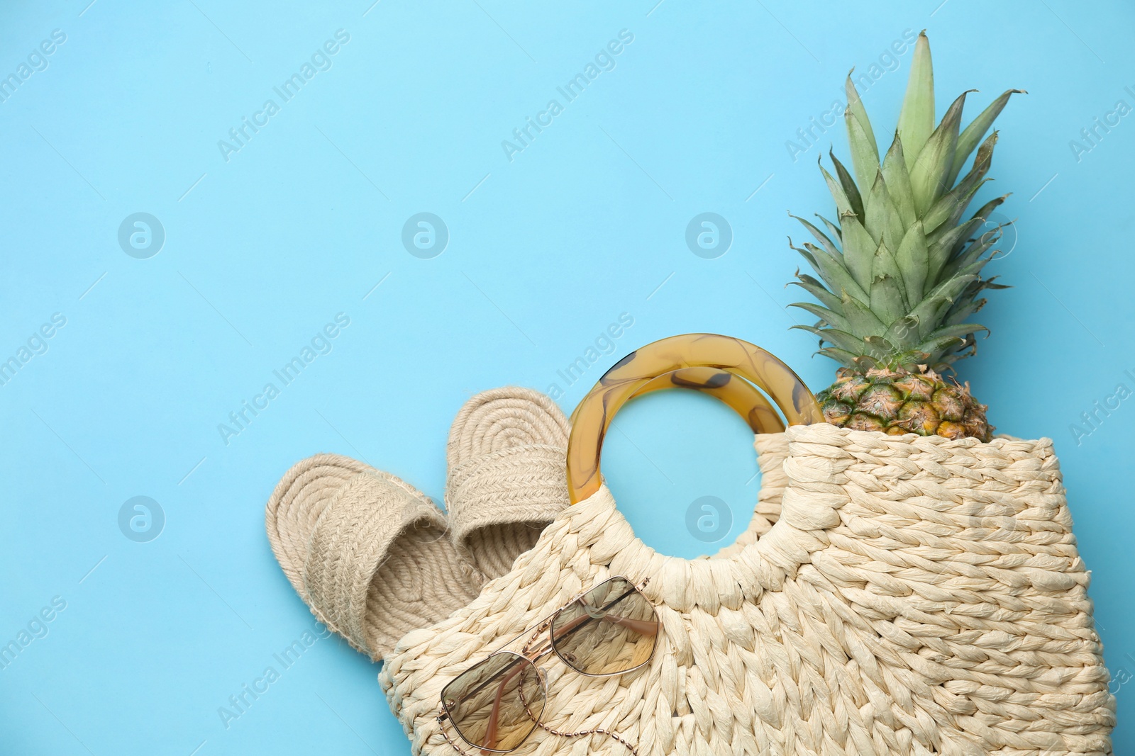 Photo of Elegant woman's straw bag with shoes, sunglasses and pineapple on light blue background, top view. Space for text