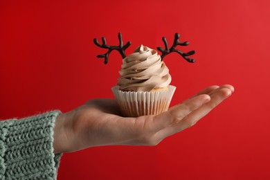 Photo of Woman holding tasty Christmas cupcake with chocolate reindeer antlers on red background, closeup
