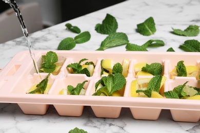 Pouring water into ice cube tray with mint and lemon on marble table