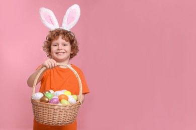 Portrait of happy boy in cute bunny ears headband holding wicker basket with Easter eggs on pink background. Space for text