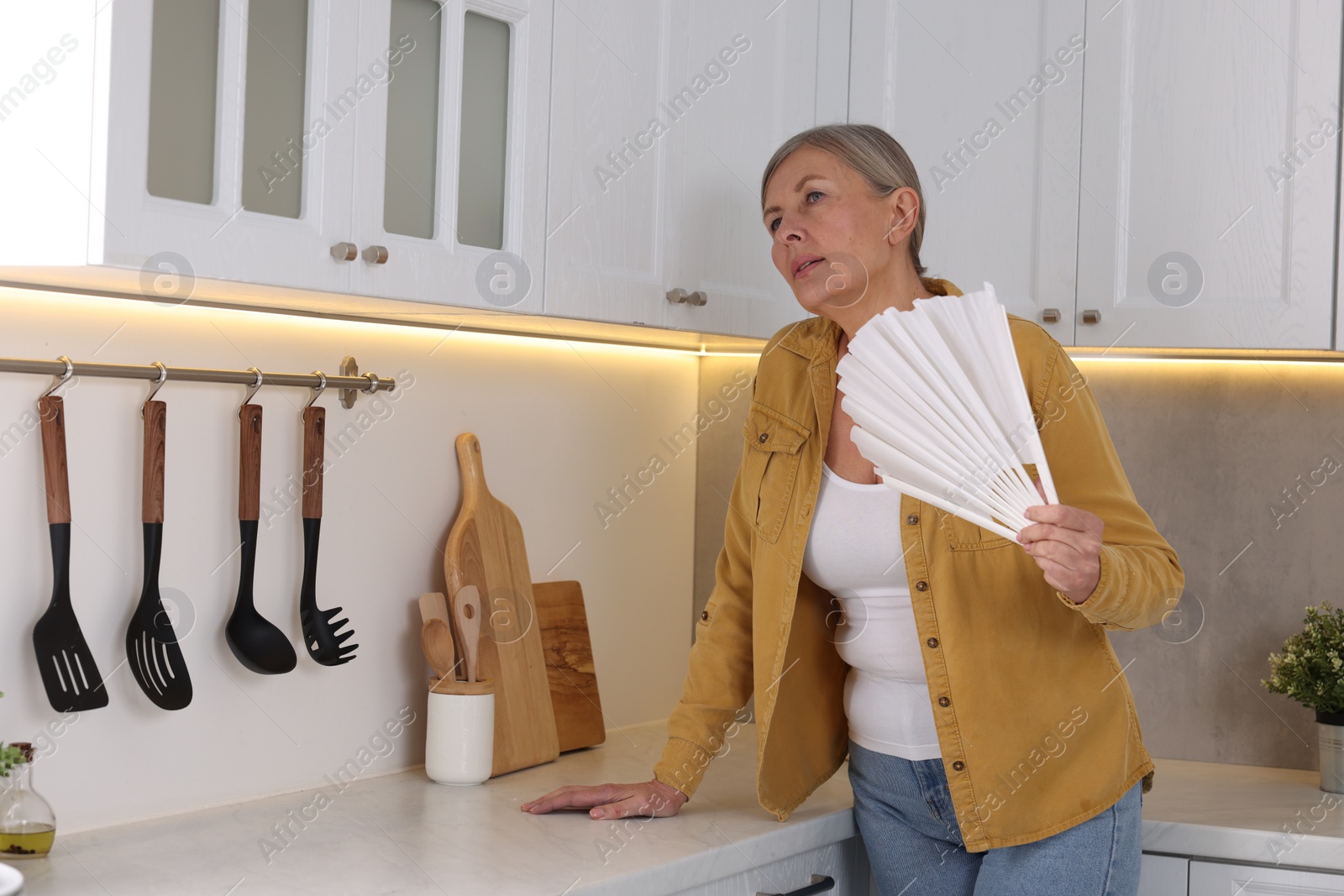 Photo of Menopause. Woman waving hand fan to cool herself during hot flash in kitchen