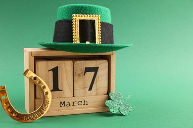 Photo of St. Patrick's day - 17th of March. Wooden block calendar, leprechaun hat, golden horseshoe and decorative clover leaf on green background. Space for text
